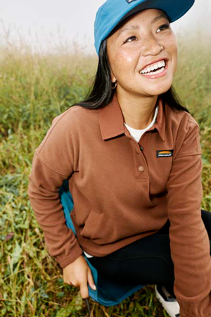 Woman sitting on a chair while camping, wearing a brown Chlorophylle fleece jacket.